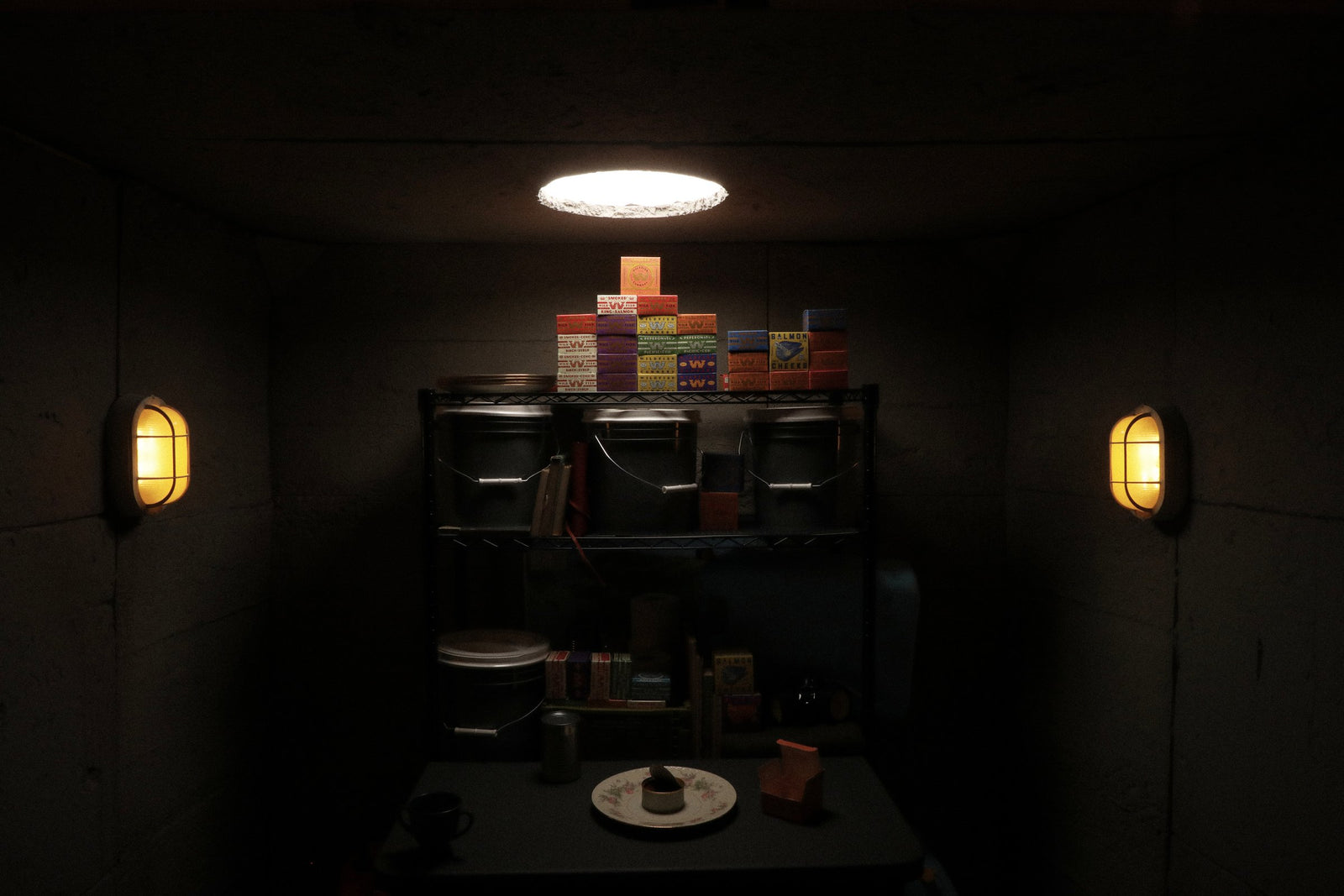 A dark concrete bunker room lit by one ceiling light and two wall lamps, a shelf of tins at the back.
