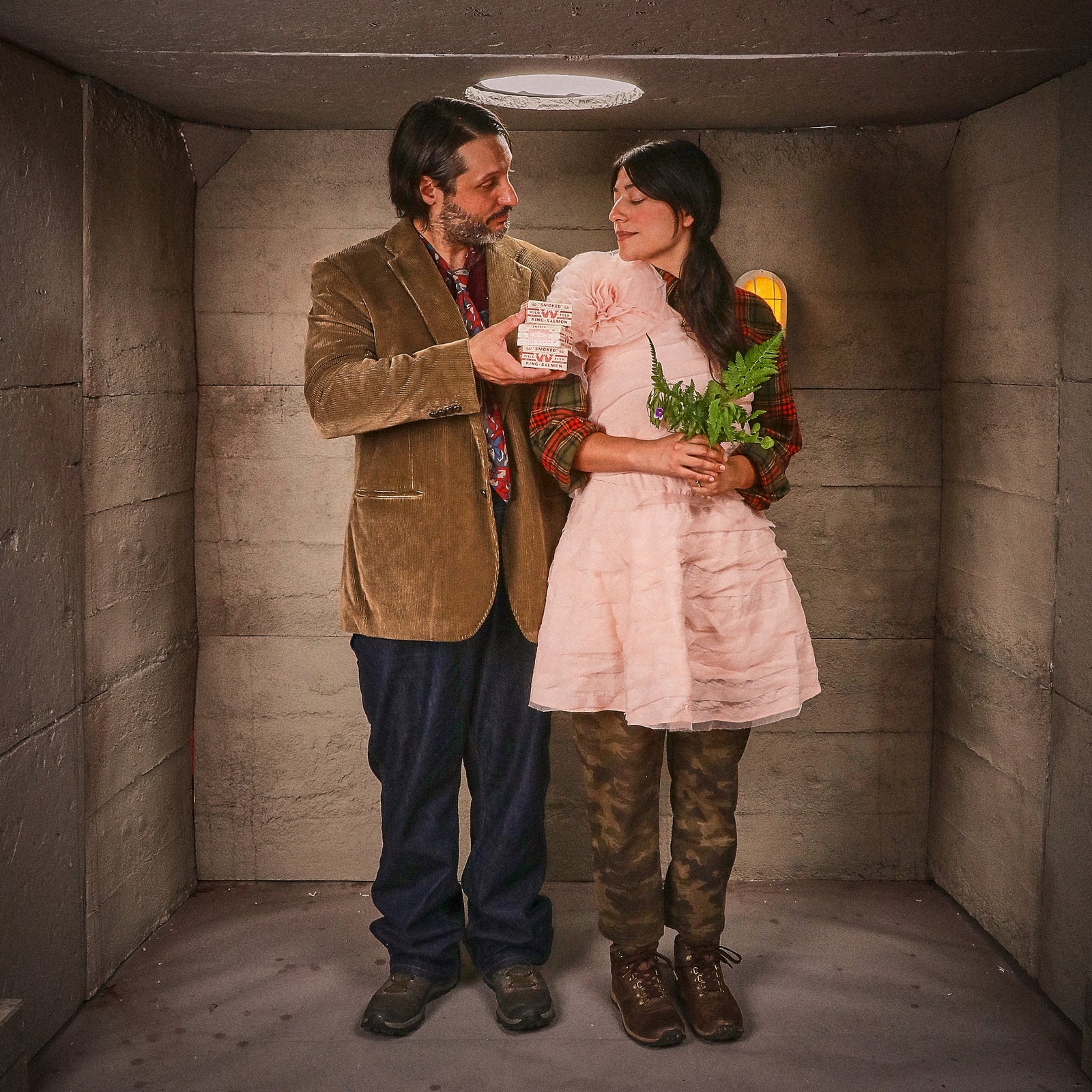 The couple stand together in the empty bunker, he in a corduroy jacket with a carton, she in a pink dress with ferns.