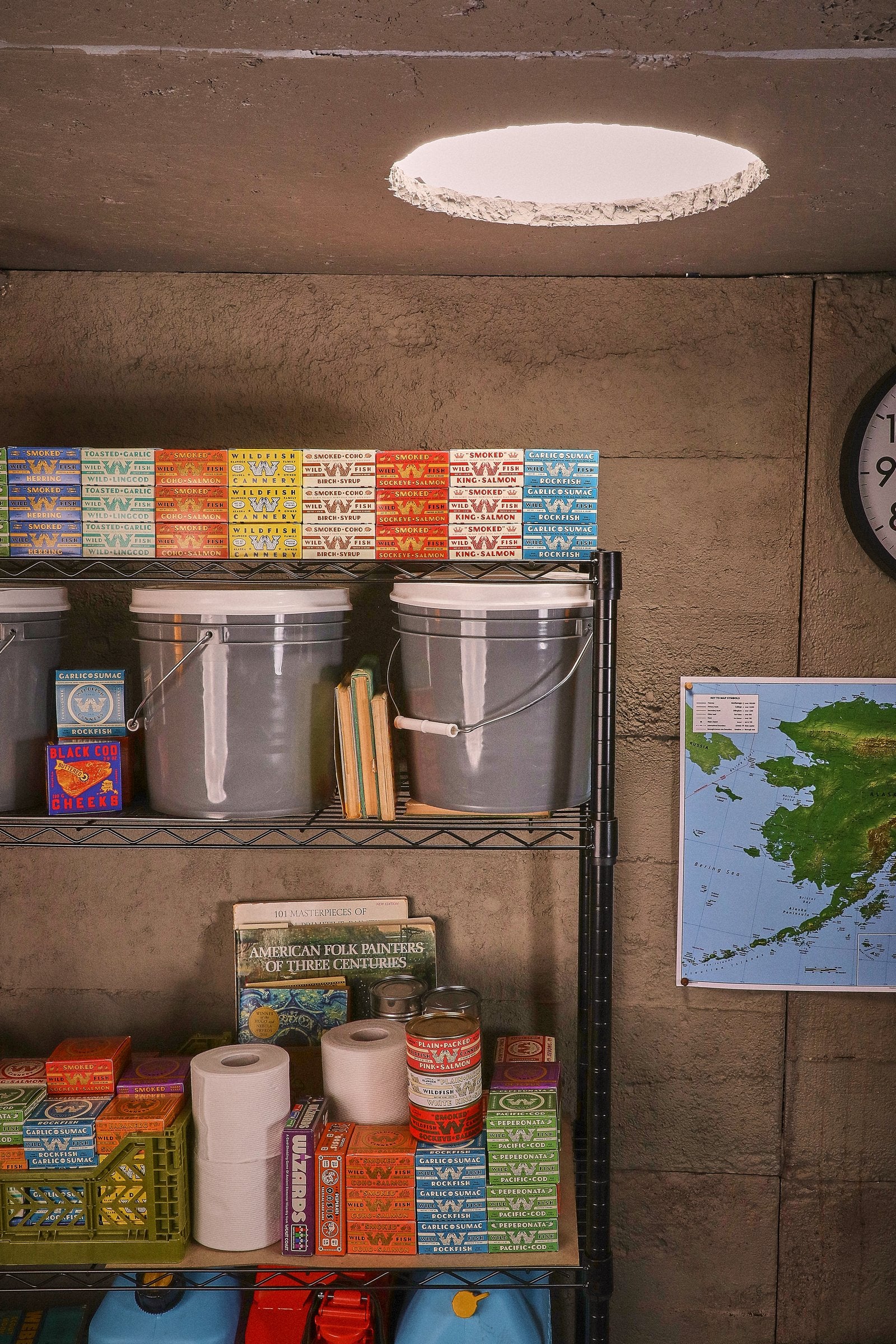 A full steel shelf under the ceiling light: cartons on top, buckets, books and a crate below, a map on the wall.