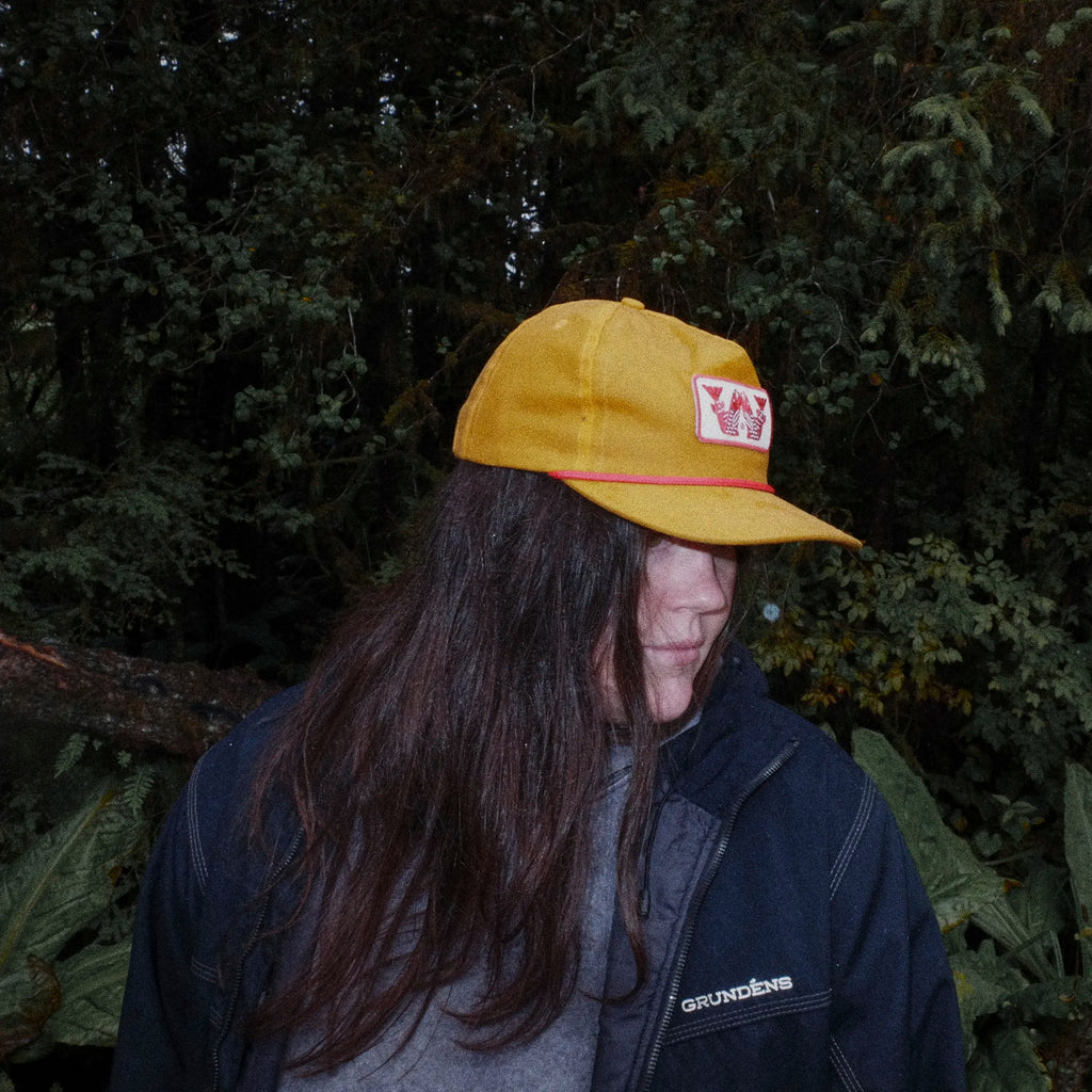 Person wearing a yellow cap with a logo, standing in front of green foliage.