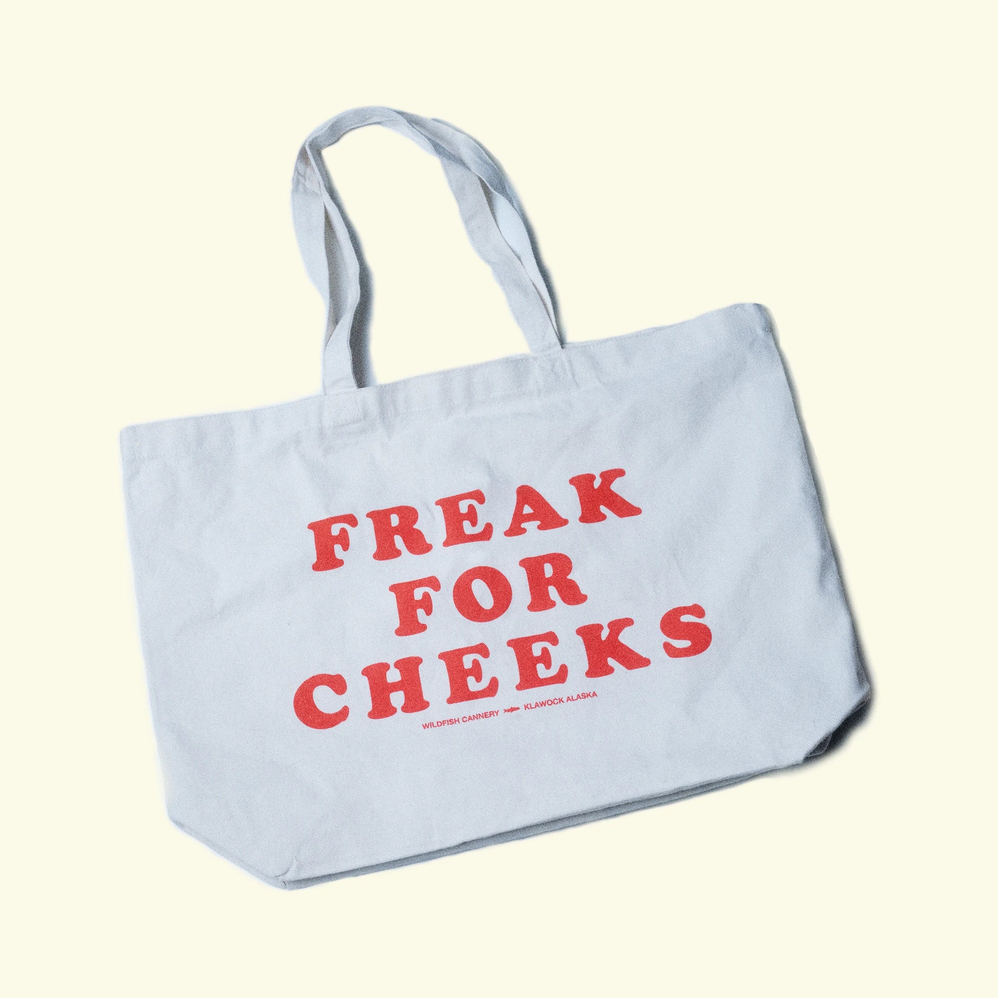 A natural canvas tote bag with the phrase 'FREAK FOR CHEEKS' printed in red across the front, set against a solid pale yellow background.