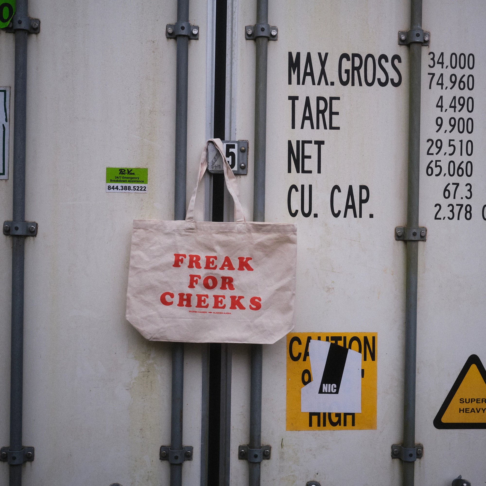 A beige canvas tote bag with red text reading 'FREAK FOR CHEEKS' hanging from a metal latch on a shipping container door. The container has black printed weight and capacity text and several warning labels.