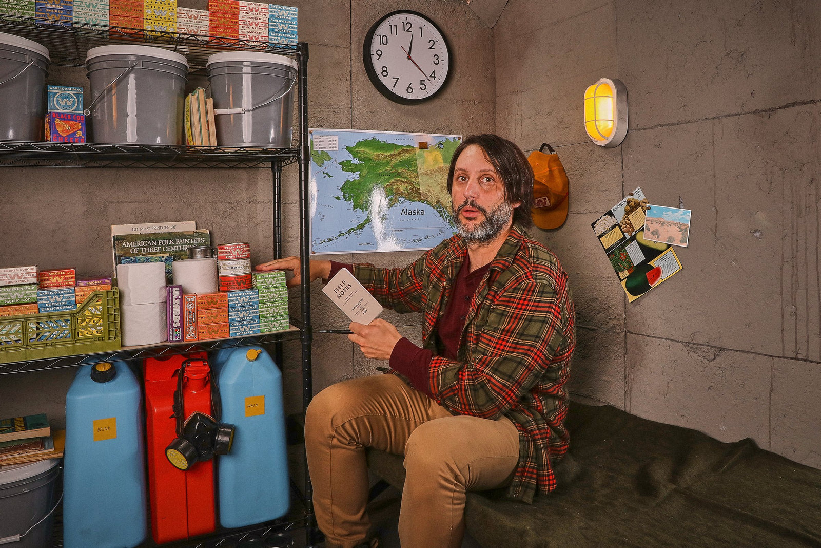 The man sits on a cot against the stocked shelves, reading a small notebook and glancing up.